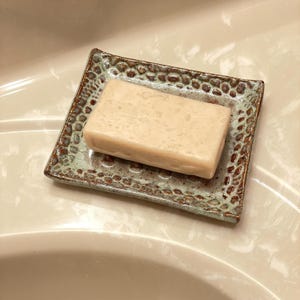 A rectangular bar of soap rests on a square ceramic soap dish. The dish has a textured, speckled design in shades of brown and green. The soap is a light beige color.