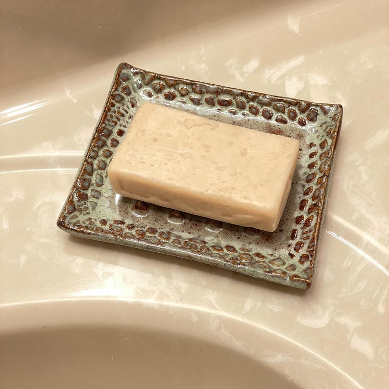 A rectangular bar of soap rests on a square ceramic soap dish. The dish has a textured, speckled design in shades of brown and green. The soap is a light beige color.
