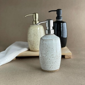 May include: Three ceramic soap dispensers with metal pumps. The dispensers are in cream, speckled white, and black. They sit on a wooden tray with a white cloth. The dispensers have a rounded shape.