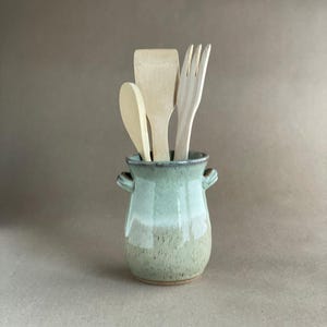 May include: A ceramic utensil holder with a light green and tan glaze, containing wooden spoons, a spatula, and a fork. The holder has small handles and is suitable for kitchen use.