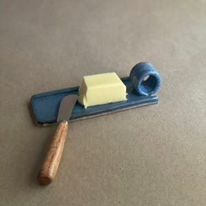 May include: A blue ceramic butter dish with a small, rectangular butter block and a butter knife with a wooden handle. The dish has a built-in ring for a thumb grip. The knife is resting on the dish.