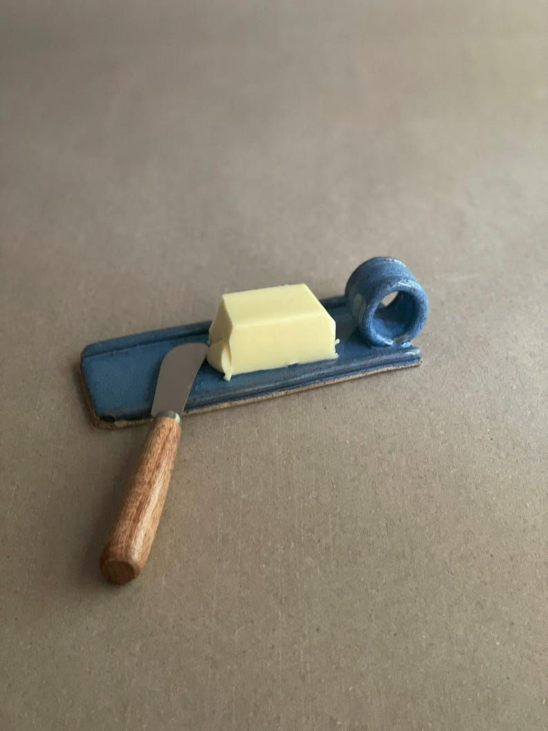 May include: A blue ceramic butter dish with a small, rectangular shape, a built-in handle, and a butter knife. The butter is a light yellow colour. The knife has a wooden handle and a silver blade.