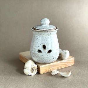 May include: A white ceramic garlic keeper with a lid, speckled with brown spots, and three decorative cutouts. The keeper sits on a small wooden block, with garlic bulbs and cloves nearby. The keeper is designed for kitchen storage.