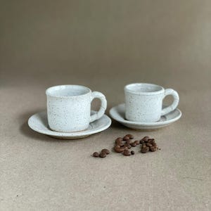 Pode incluir: Duas pequenas chávenas de café expresso de cerâmica branca salpicada com pires a condizer. As chávenas têm pegas curvas e são acompanhadas por uma pequena pilha de grãos de café.