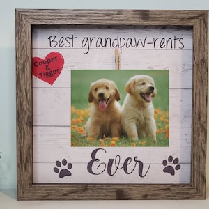 May include: A wooden picture frame with a white background and a photo of two golden retriever puppies. The frame has the text "Best grandpaw-rents" at the top, a red heart with the text "Cooper & Tigger" on the left, and the text "Ever" with paw prints on the bottom.