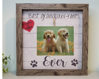 May include: A wooden picture frame with a white background and a photo of two golden retriever puppies. The frame has the text "Best grandpaw-rents" at the top, a red heart with the text "Cooper & Tigger" on the left, and the text "Ever" with paw prints on the bottom.