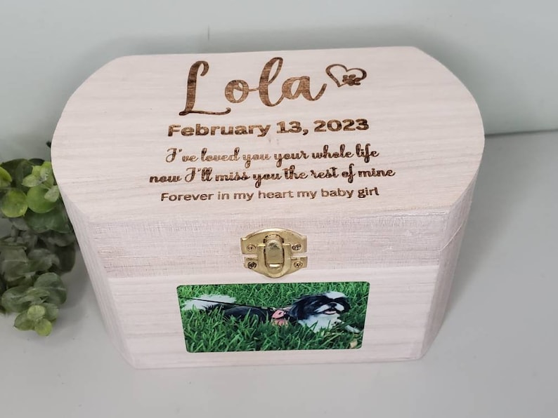K&ouml;nnte beinhalten: Eine wei&szlig;e Holzkiste mit einem Scharnierdeckel und einem Messingverschluss. Die Kiste hat ein Foto eines Hundes auf der Vorderseite und ist mit dem Text "Lola", "13. Februar 2023", "Ich habe dich dein ganzes Leben lang geliebt, jetzt werde ich dich f&uuml;r den Rest meines Lebens vermissen" und "F&uuml;r immer in meinem Herzen mein kleines M&auml;dchen" graviert.