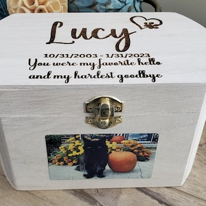 K&ouml;nnte beinhalten: Eine wei&szlig;e Holzkiste mit einem Scharnierdeckel und einem Messingverschluss. Der Deckel ist mit dem Text "Lucy 10/31/2003 - 1/31/2023 You were my favorite hello and my hardest goodbye" graviert. Ein Foto einer schwarzen Katze ist auf der Vorderseite der Kiste angebracht.