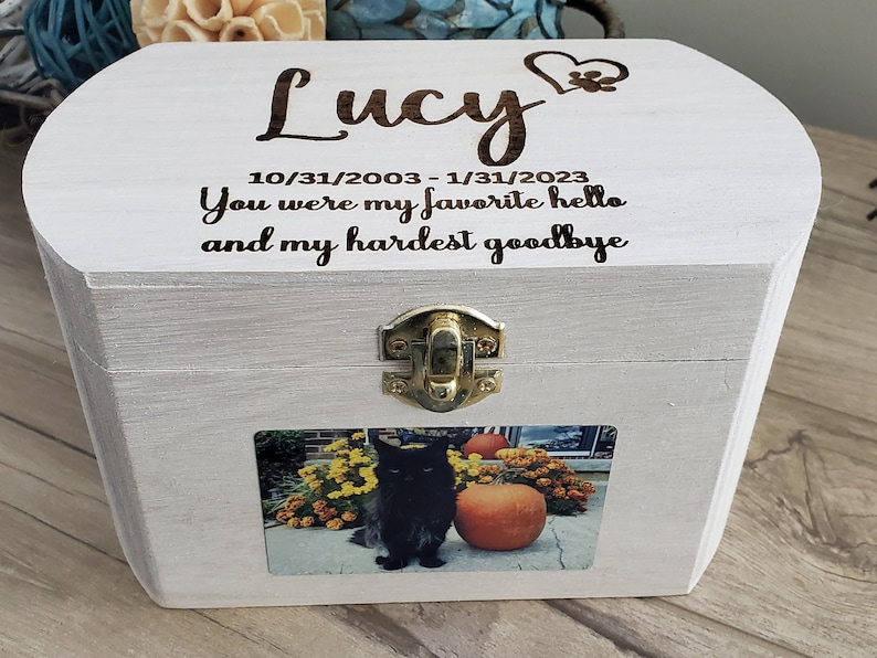 K&ouml;nnte beinhalten: Eine wei&szlig;e Holzkiste mit einem Scharnierdeckel und einem Messingverschluss. Der Deckel ist mit dem Text "Lucy 10/31/2003 - 1/31/2023 You were my favorite hello and my hardest goodbye" graviert. Ein Foto einer schwarzen Katze ist auf der Vorderseite der Kiste angebracht.