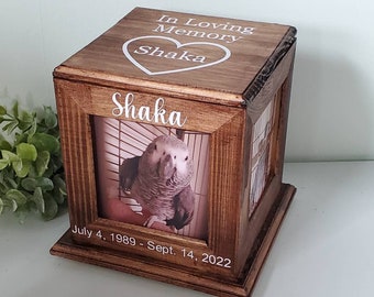 May include: Wooden memorial box with a photo of a grey parrot in a cage. The box has a heart with the name "Shaka" and the text "In Loving Memory" on the top. The bottom of the box has the text "July 4, 1989 - Sept. 14, 2022" and the name "Shaka" on the front.