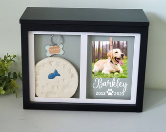 Puede incluir: Caja conmemorativa para mascotas enmarcada en negro con dos compartimentos. Un lado presenta una foto de un Labrador Retriever amarillo con el nombre "Barkley" y las fechas 2013-2023. El otro lado tiene una huella de pata y una placa de identificación para perros.