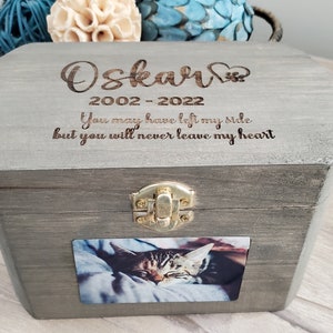 K&ouml;nnte beinhalten: Eine graue Holzkiste mit einem Scharnierdeckel und einem goldenen Verschluss. Die Kiste hat ein Foto einer Katze auf der Vorderseite und ist mit dem Text "Oskar 2002-2022 You may have left my side but you will never leave my heart" graviert.