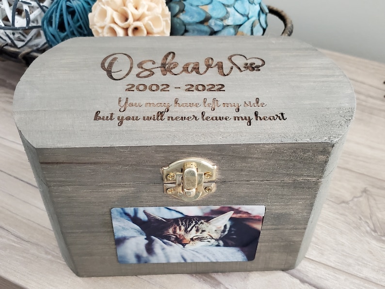 K&ouml;nnte beinhalten: Eine graue Holzkiste mit einem Scharnierdeckel und einem goldenen Verschluss. Die Kiste hat ein Foto einer Katze auf der Vorderseite und ist mit dem Text "Oskar 2002-2022 You may have left my side but you will never leave my heart" graviert.