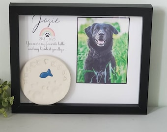 May include: A black framed picture with a photo of a black dog in a field of green grass. The picture has a white mat and a white background with the text "Josie 2013-2023 You were my favorite hello and my hardest goodbye". There is a white ceramic paw print with a blue fish on it in the lower left corner of the picture.
