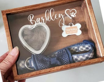 May include: A wooden shadow box with a glass front, containing a dog's collar, a bow tie, a heart-shaped container with fur, and a bone-shaped tag with the name "Barkley" on it. The box is personalized with the name "Barkley" and a paw print.