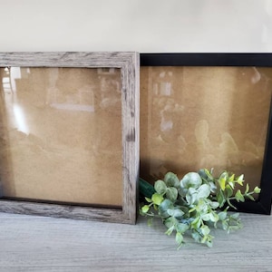 Puede incluir: Dos marcos de fotos, uno con un acabado de madera gris y otro con un acabado negro. Ambos marcos tienen un frente de vidrio transparente y un respaldo marr&oacute;n.
