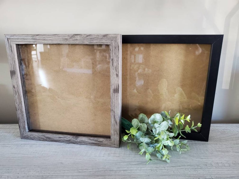 Puede incluir: Dos marcos de fotos, uno con un acabado de madera gris y otro con un acabado negro. Ambos marcos tienen un frente de vidrio transparente y un respaldo marr&oacute;n.