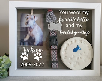 May include: A white shadow box memorial for a pet with a photo of a white dog, a paw print clay imprint, a collar, and the text "You were my favorite hello and my hardest goodbye", "Jackson", and "2009-2022".