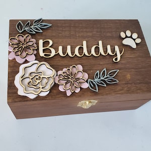 May include: A wooden box with a hinged lid and a brass latch. The box is decorated with a floral design and the name "Buddy" in cursive. A paw print is also visible on the box.