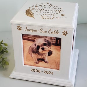 May include: White wooden box with a photo of a small dog inside. The box has a gold inscription that reads "You may have left my life but you will never leave my heart." The inscription also includes paw prints and a feather. The box has a name plaque that reads "Jacque-Sue Cable" and dates "2008-2023".