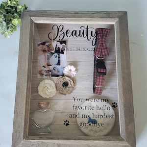 May include: A wooden shadow box memorial for a dog named Beauty. The box includes photos of the dog, a red and black plaid dog collar, a small glass jar, a white flower, a brown burlap flower, a white ceramic paw print with the text "You were my favorite hello and my hardest goodbye", and the dates "01-01-2005 - 04-16-2014".