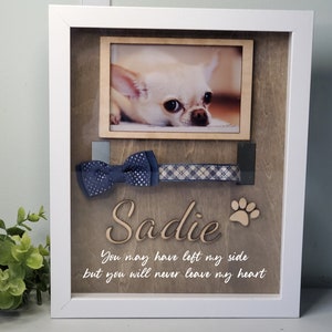 May include: A white wooden shadow box with a photo of a small white dog, a blue bow tie, a plaid collar, and the name "Sadie" written in wood. The text "You may have left my side but you will never leave my heart" is written below the name.