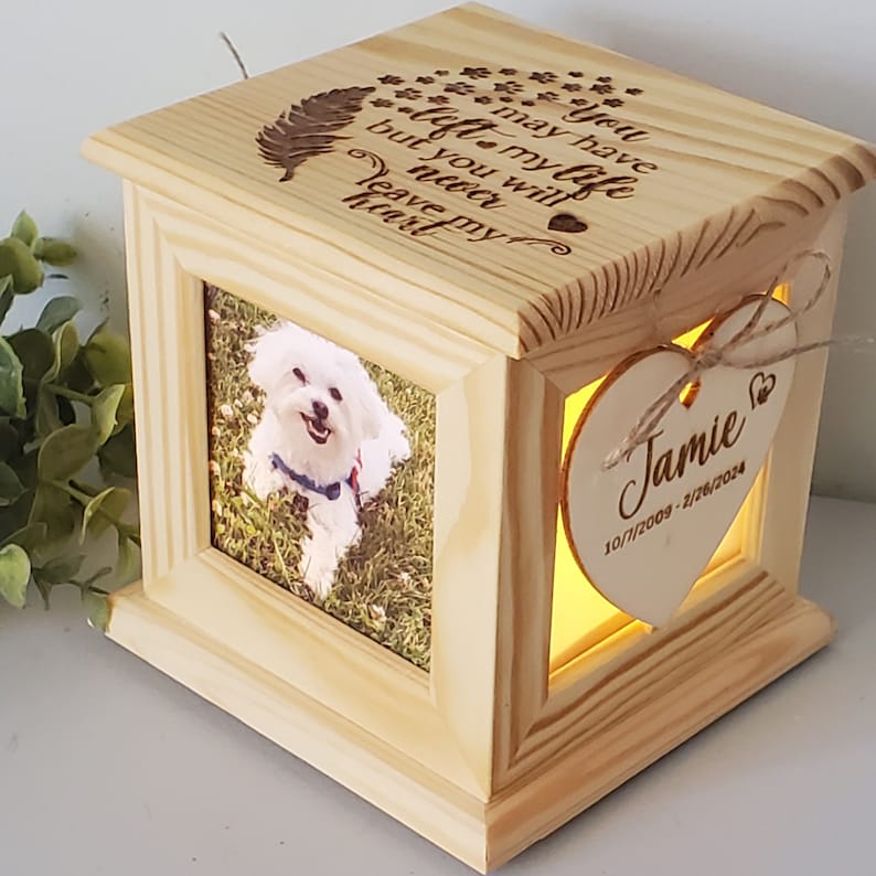 Puede incluir: Una caja de madera conmemorativa con una foto de un perro blanco. La caja tiene una luz en el interior y una placa en forma de coraz&oacute;n con la inscripci&oacute;n "You may have left my life but you will never leave my heart" y el nombre "Jamie" con fechas.