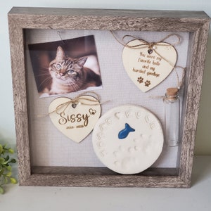 May include: A wooden shadow box memorial with a photo of a cat, a paw print, a small glass vial, and two wooden hearts with the text "Sissy 2013-2023" and "You were my favorite hello and my hardest goodbye".