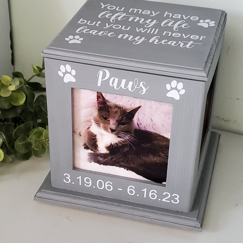 Może przedstawiać: Szare drewniane pudełko z fotografią kota w środku. Pudełko ma napis "You may have left my life but you will never leave my heart" i "Paws". Daty "3.19.06 - 6.16.23" r&oacute;wnież znajdują się na pudełku.