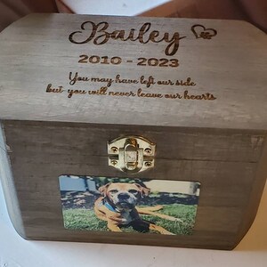 K&ouml;nnte beinhalten: Eine Holzkiste mit einem Scharnierdeckel und einem Messingverschluss. Die Kiste ist mit dem Namen "Bailey" und den Daten "2010-2023" graviert. Die Inschrift lautet "You may have left our side but you will never leave our hearts". Ein Foto eines Hundes ist an der Vorderseite der Kiste befestigt.