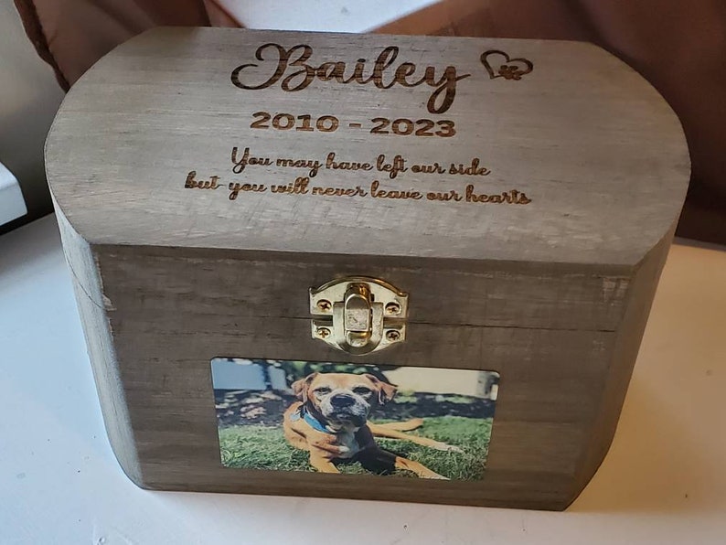 K&ouml;nnte beinhalten: Eine Holzkiste mit einem Scharnierdeckel und einem Messingverschluss. Die Kiste ist mit dem Namen "Bailey" und den Daten "2010-2023" graviert. Die Inschrift lautet "You may have left our side but you will never leave our hearts". Ein Foto eines Hundes ist an der Vorderseite der Kiste befestigt.
