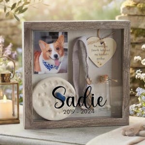 May include: A square wooden shadow box frame containing a photograph of a corgi, a grey fabric collar, a heart-shaped wooden tag with the text 'You were my favorite hello and my hardest goodbye', and a clay paw print. The glass features the name 'Sadie' and '2019 - 2024'.