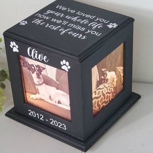 May include: Black wooden box with a photo of a dog inside. The box has a white inscription that reads "We've loved you your whole life now we'll miss you the rest of ours" and "Clive" with paw prints. The bottom of the box has the dates "2012-2023" inscribed on it.