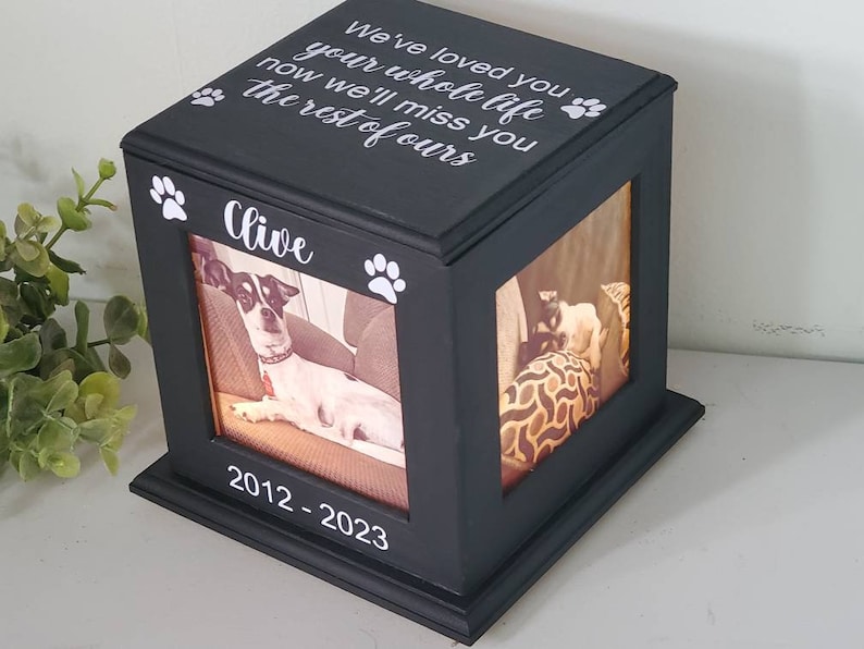 May include: Black wooden box with a photo of a dog inside. The box has a white inscription that reads "We've loved you your whole life now we'll miss you the rest of ours" and "Clive" with paw prints. The bottom of the box has the dates "2012-2023" inscribed on it.