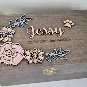 May include: A wooden box with a hinged lid, decorated with laser-cut flowers, leaves, and the name "Jessy". The box features a paw print and the dates 03/10/2005-06/06/2025. The box has a gold-colored clasp.
