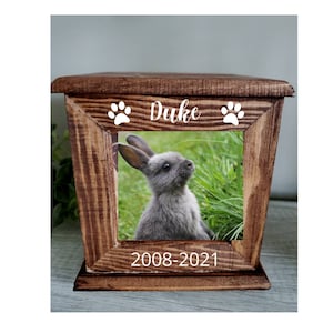 May include: A rustic wooden pet memorial box with a framed photo of a gray rabbit. The box is engraved with the name "Duke", paw prints, and the years "2008-2021". The box has a natural wood grain finish.