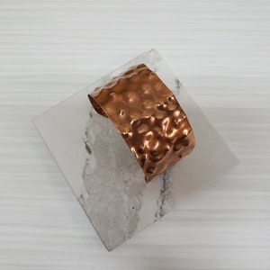 Puede incluir: Un brazalete de puño de cobre martillado con una superficie texturizada. El brazalete ancho tiene un color marrón rojizo cálido y una forma ligeramente curvada. El brazalete se exhibe sobre una superficie cuadrada marmolada.