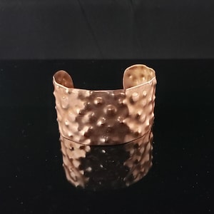 Puede incluir: Un brazalete de color cobre con una superficie texturizada y martillada. El brazalete abierto tiene una banda ancha y está diseñado para usarse en la muñeca. El metal refleja la luz, creando reflejos y sombras.