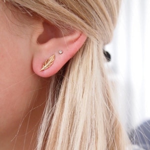 May include: A close-up of a person's ear with two small earrings. The earring on the lower lobe is a gold feather design. The earring on the upper lobe is a small, clear diamond.