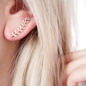 Może przedstawiać: Kolczyk na ucho w kolorze różowego złota w kształcie liścia, ozdobiony małymi, błyszczącymi kryształkami. Kolczyk zakrzywia się wzdłuż ucha, tworząc delikatny i elegancki wygląd. Zdjęcie przedstawia kolczyk na osobie z blond włosami.