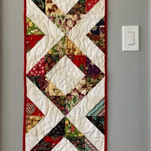 May include: A rectangular quilted wall hanging with a red border. The design features white diamond shapes with colourful fabric inserts. The inserts include patterns with red, green, and gold hues. The quilt is hanging on a grey wall.