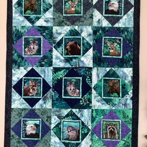 May include: A green, blue, and purple quilt with a black border. The quilt is made up of 12 squares, each with a different animal image in the center. The animals include a moose, deer, eagle, wolf, bear, raccoon, fox, otter, and a few more.