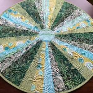 May include: A round table runner with a green, blue and yellow swirl pattern. The runner is made of fabric and has a circular shape.