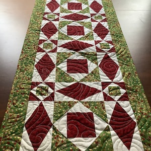 May include: A quilted table runner with a geometric pattern in shades of green, white, and burgundy. The runner features a repeating design of squares, diamonds, and swirls.