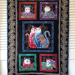 May include: A black quilted wall hanging with a colorful paisley design featuring cats. The cats are in various poses and have different colored eyes. The wall hanging is framed with a decorative border of gold and silver.