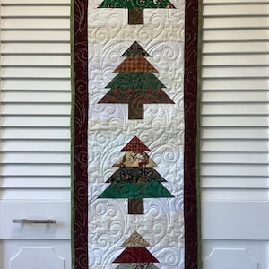 May include: A quilted wall hanging featuring a festive Christmas design. Four patchwork trees are arranged vertically, each with a different pattern of red, green and white fabrics. The trees are set against a white background with a burgundy and green border.