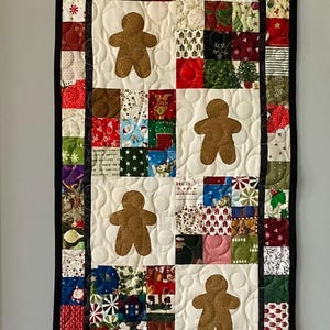 Peut inclure: Chemin de table matelassé sur le thème de Noël. Le centre est blanc avec des appliqués de bonhommes en pain d'épices bruns. Les bordures sont faites de divers tissus de Noël rouges, verts et blancs. Le chemin de table mesure environ 30 cm de large et 91 cm de long.