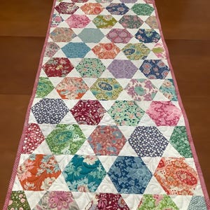 May include: A handmade table runner with a patchwork design. It features hexagonal fabric pieces with floral patterns in pink, blue, green, and orange. The runner is bordered with a pink and white striped fabric. The runner's length is approximately 150 cm.