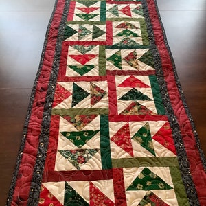 May include: A quilted table runner with a Christmas theme. The runner is made of red, green, and gold fabric and features a repeating pattern of triangles. The runner is edged with a black and gold trim.