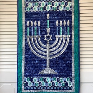 May include: A blue and white quilted wall hanging featuring a silver menorah with lit candles. The background is filled with Hebrew text and the words "Lights miracle Family Festival of Lights ONS hanukkah TRADITIONS hanu".